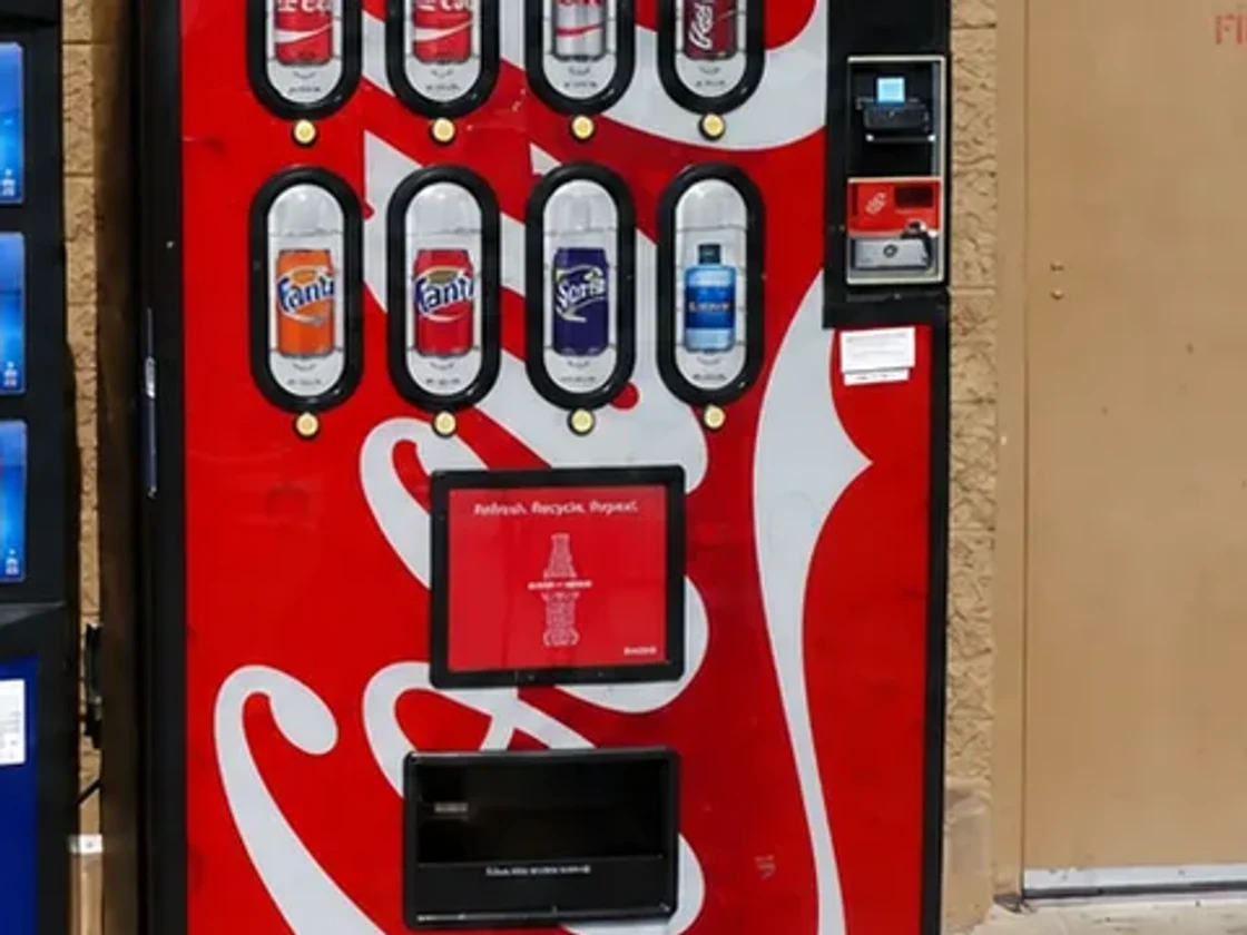 A Coca-Cola machine outdoors with a screen mounted on its front panel below the selections, running a Coca-Cola advertisement.