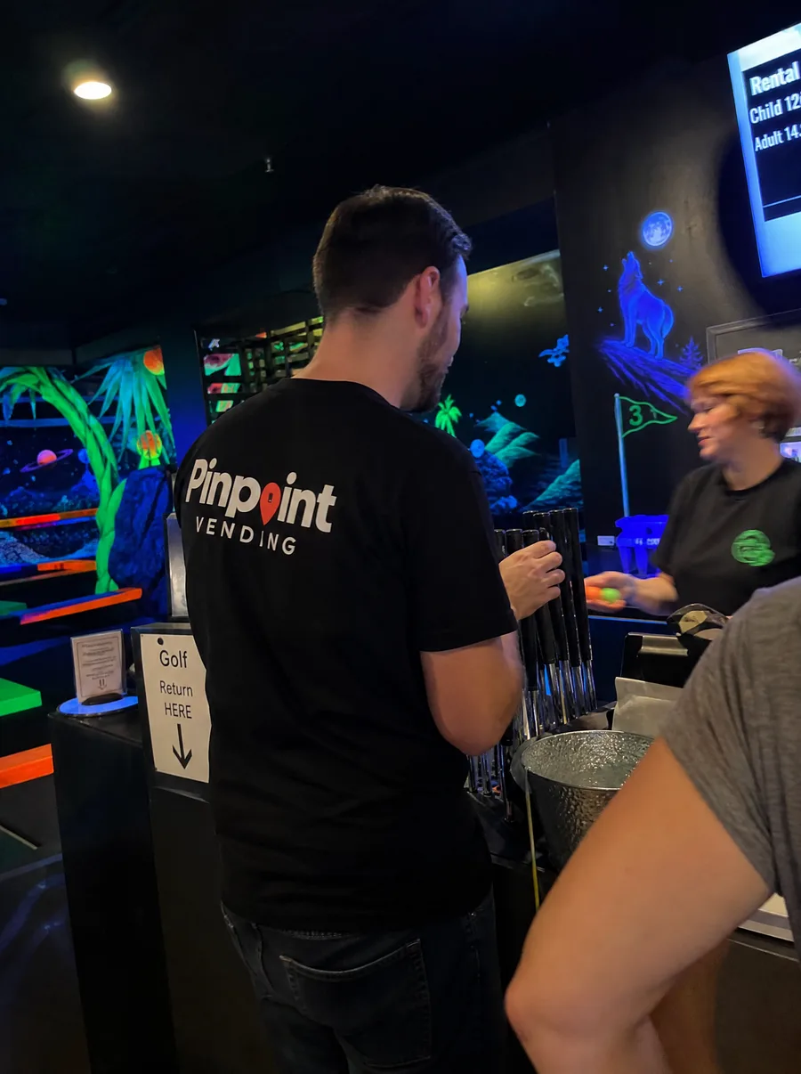 A Pinpoint rep in a branded tee at the club counter of a blacklight indoor mini golf course, a rack of putters in front of him and a member of staff alongside.