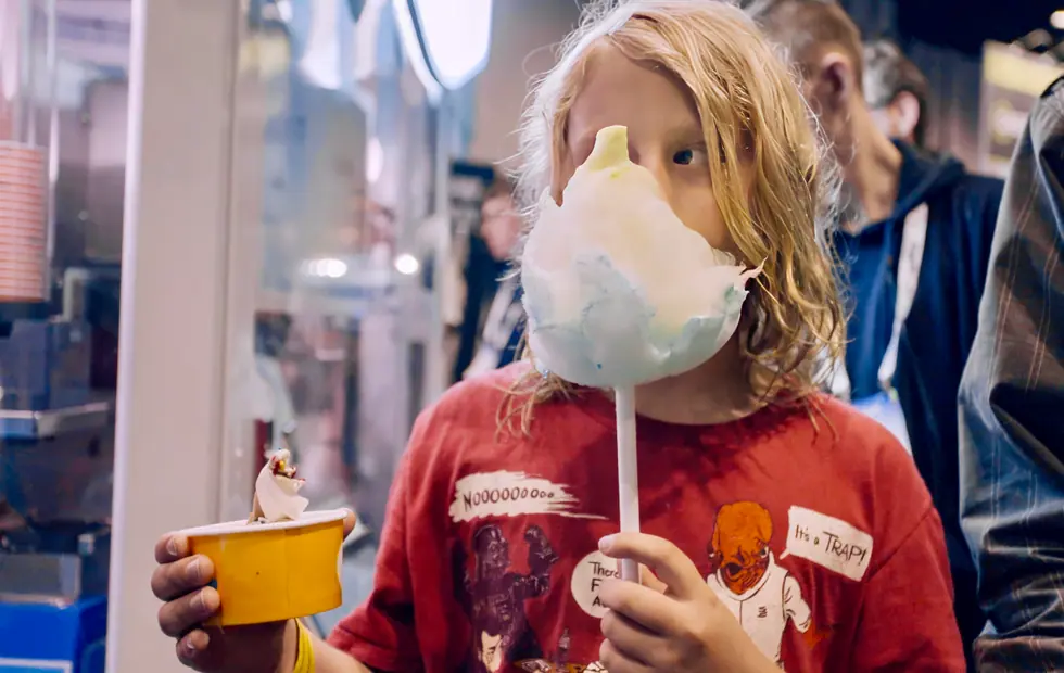 A child holding a large cotton candy up to their face, a cup in the other hand, in a busy venue.