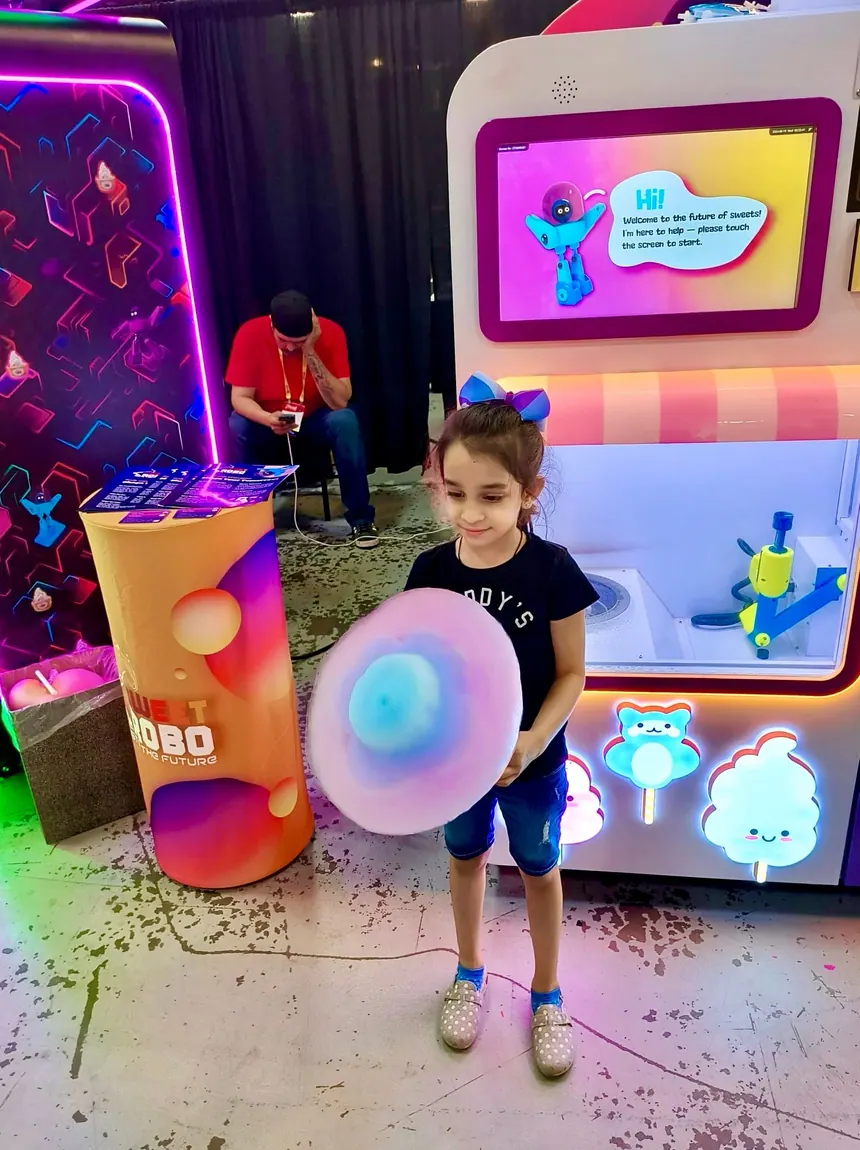 A boy holding a finished cotton candy in front of the machine, under arcade neon.