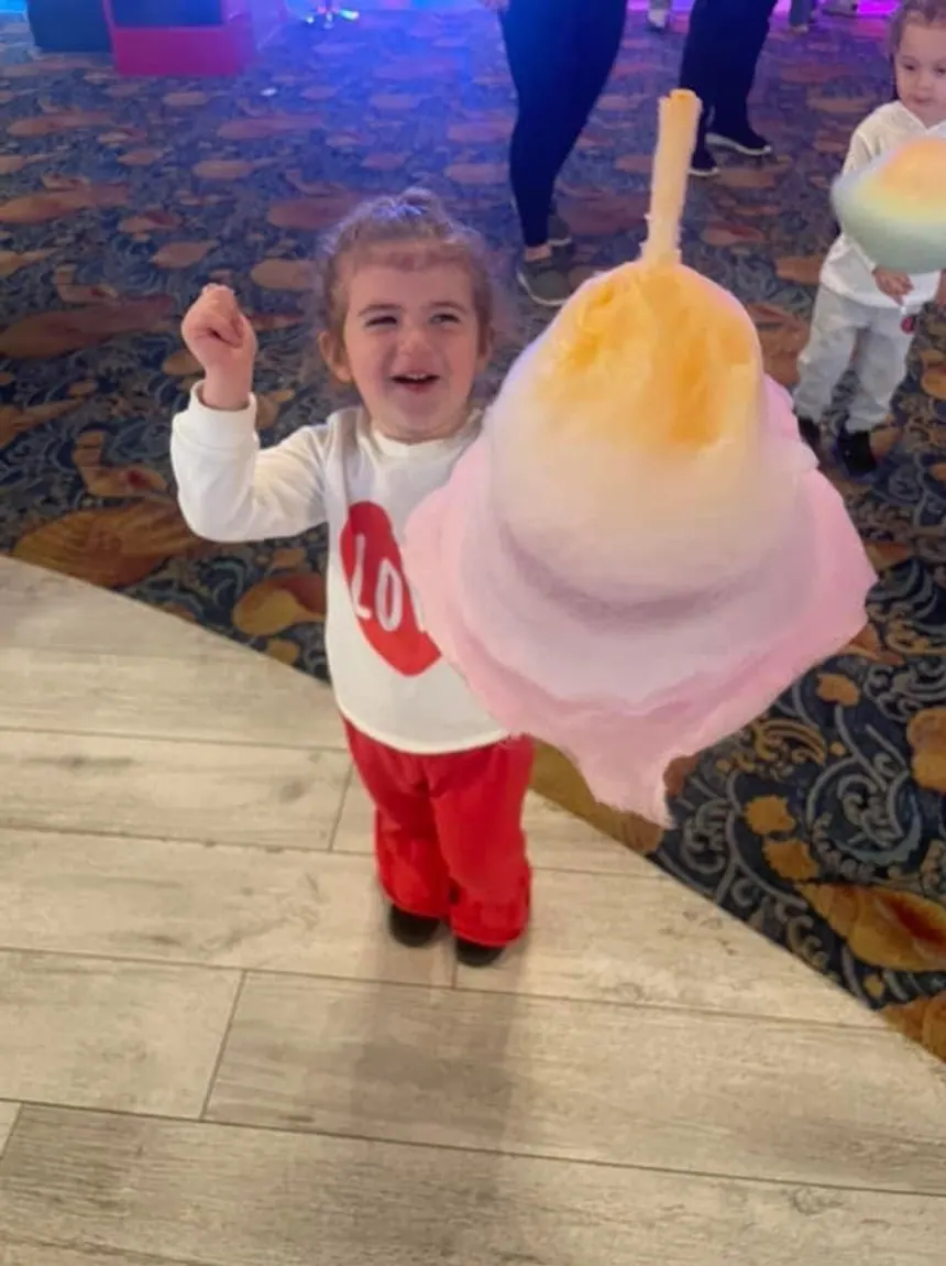 A toddler running across a patterned venue carpet with a cotton candy twice her size.