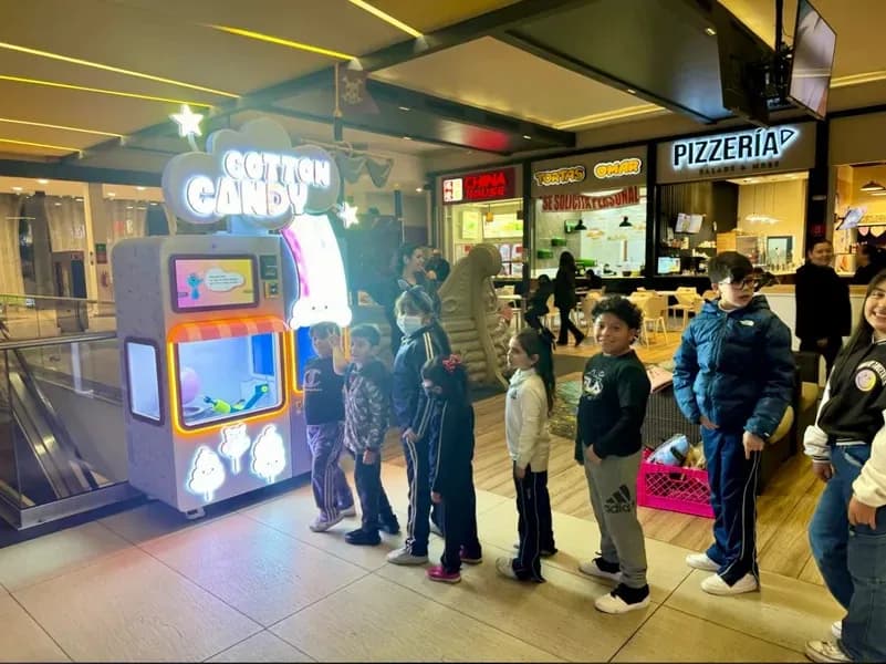 A queue of children waiting at a cotton candy vending machine in a mall food court.