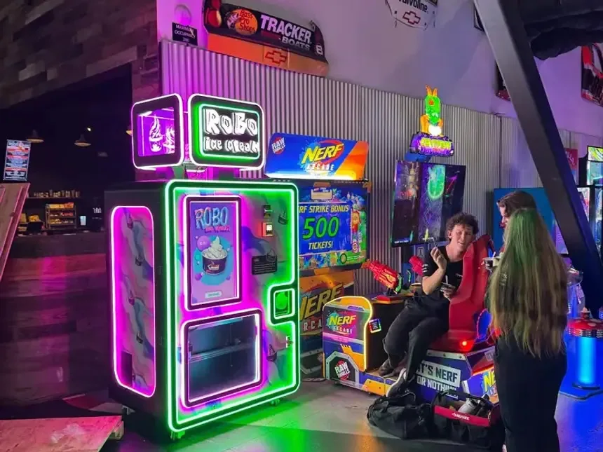 A neon-lit Robo Ice Cream vending machine on an arcade floor, with Nerf arcade cabinets and a racing simulator seat beside it.