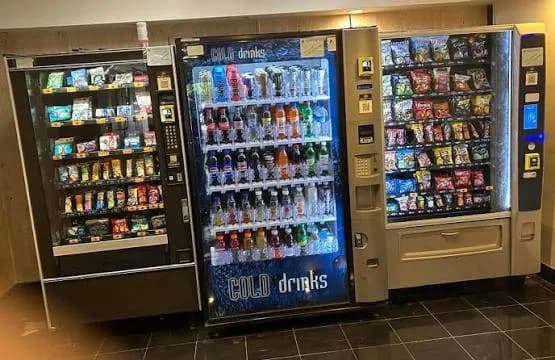 A bank of three snack and drink vending machines standing side by side against a panelled wall indoors.