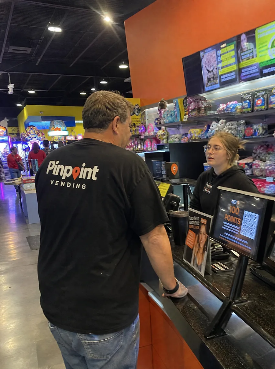 A man in a Pinpoint Vending shirt talks with a staff member across an arcade prize counter, with shelves of plush toys and prizes behind her and game cabinets in the background.