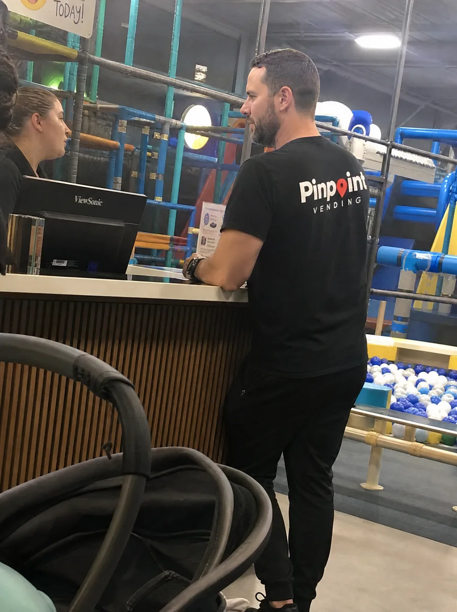 A Pinpoint rep in a branded tee at the check-in counter of an indoor playground, talking to a staff member with the climbing frame behind.