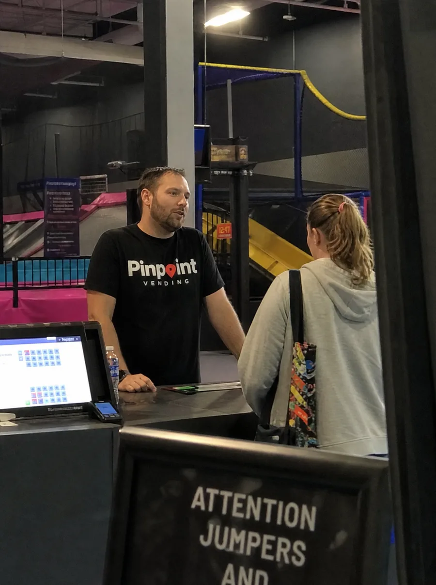 A Pinpoint rep behind the ticket counter of a trampoline park in conversation with a member of staff, jump lanes and safety netting behind them.