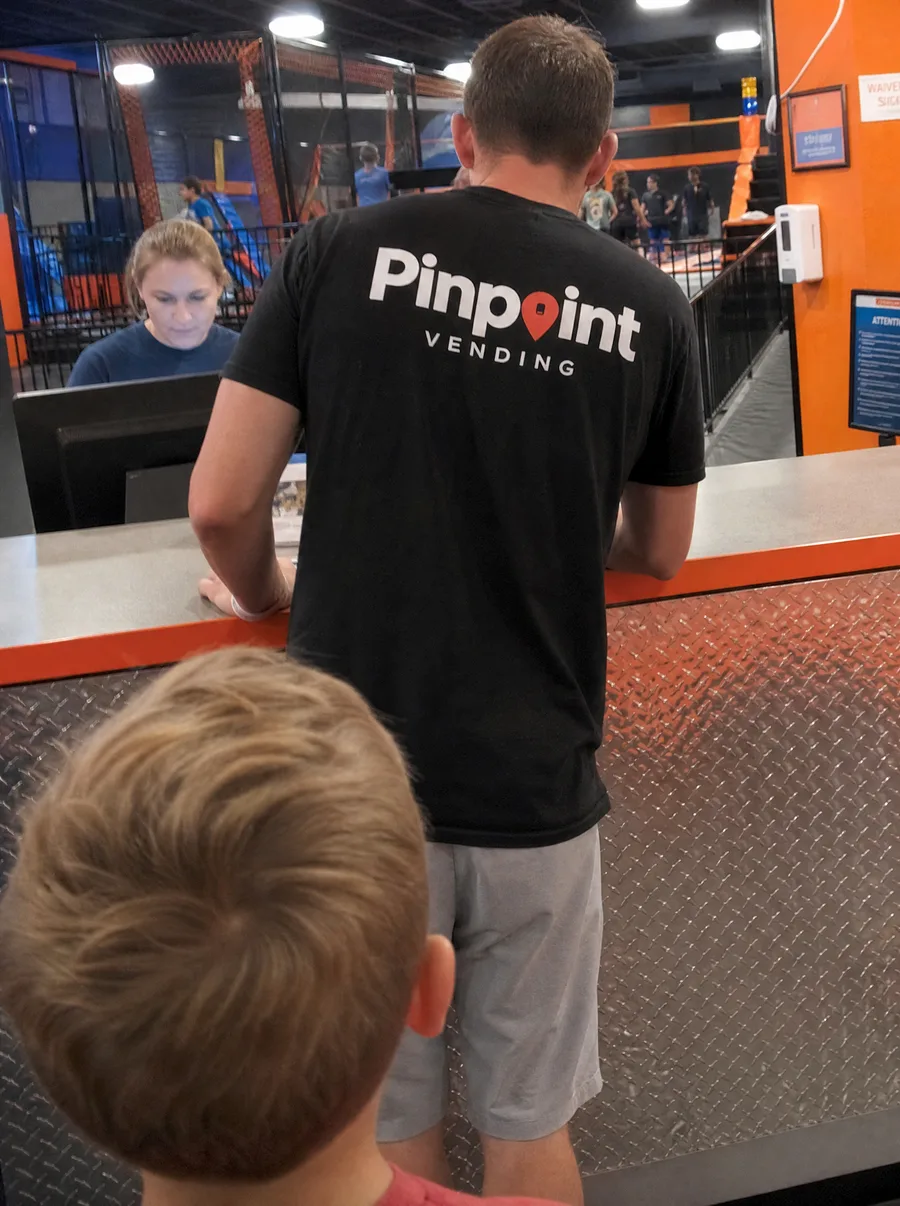 A Pinpoint rep, seen from behind in a branded tee, leaning on the waiver desk of a trampoline park while a staff member works at the computer.