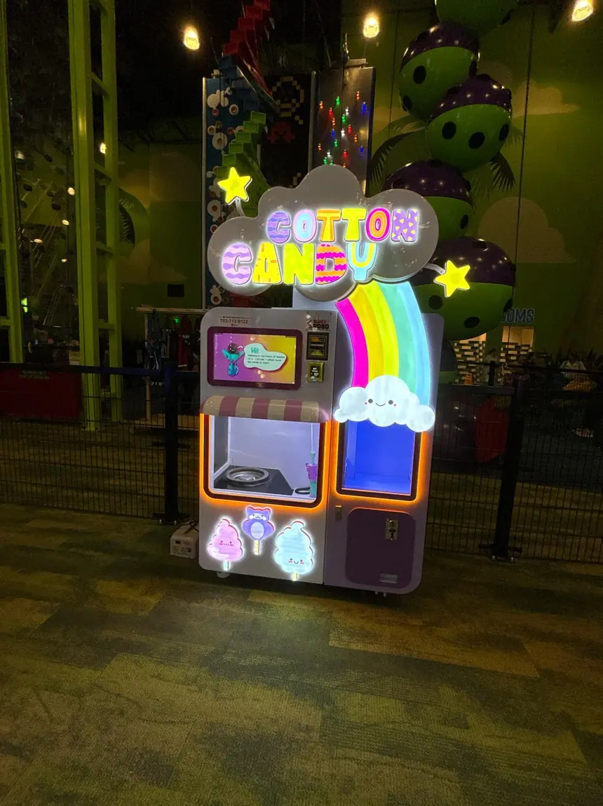 A lit cotton candy vending machine standing on carpet tile in a bowling alley, with oversized bowling-ball decor and the lanes behind it.