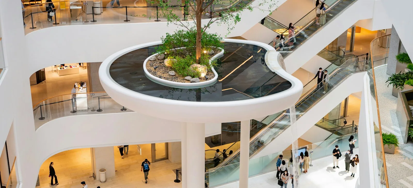 A multi-level shopping mall atrium with escalators, a planted island and shoppers on every floor.